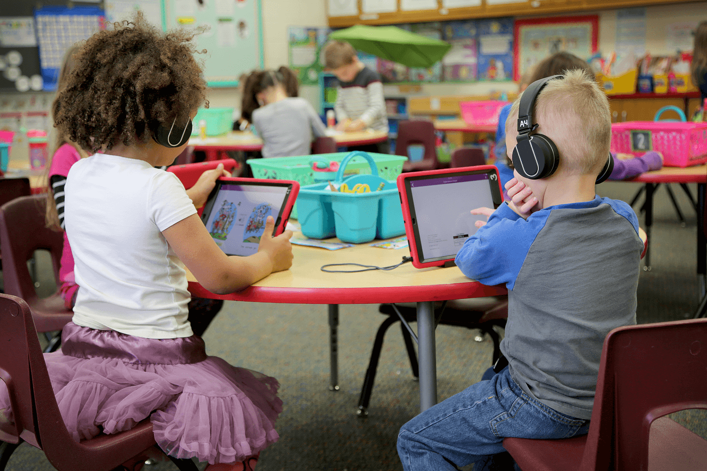 A classroom full of students sitting in chairs at their desks looking towards the front of the room at a display screen while the teacher is talking to them from the front of the room.