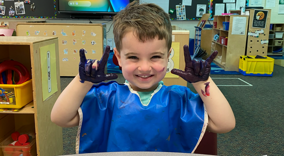 A young boy with a blue smock on holding his hands up. His hands are covered in blue paint and he's making the ASL sign for 'I love you' with both hands and smiling at the camera.