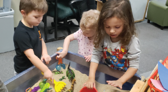 Two young girls and a young boy playing with toys in a sand table.