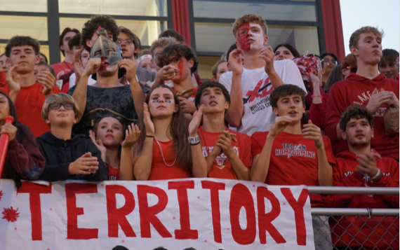 A male student with glasses and a red bandana on his head wearing a pink shirt and smiling while playing the drums.