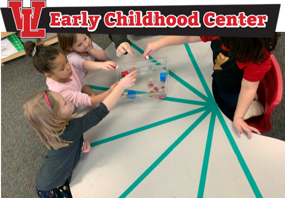 Four young students sitting at one side of a table and one young student sitting at the opposite side of the table. All of the students are placing colored items into a bin.