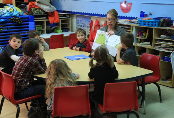 A teacher sits at the center of a table in the classroom while seven young boys and girls sit around the table facing the teacher. The teacher is holding up a piece of paper and talking to the students.