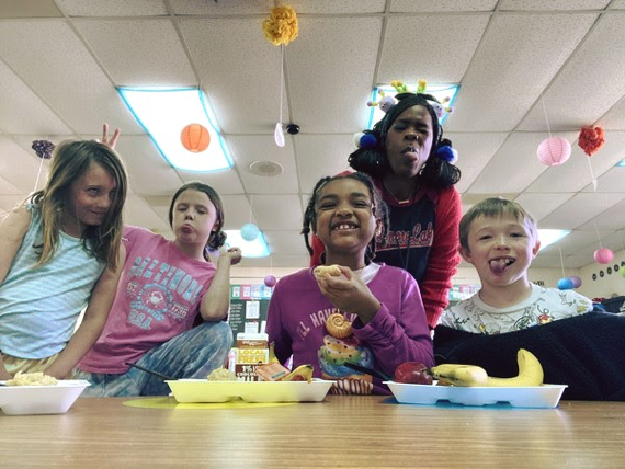 Two young male students and a young female students sit in a row at their desks. Their school work is on their desks and they are smiling at the camera.