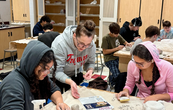 Three young girls stand in a classroom with their arms around each other while smiling at the camera.