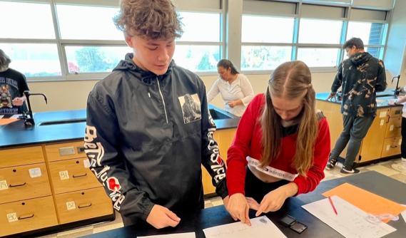 Two students performing thumb on paper in a classroom.