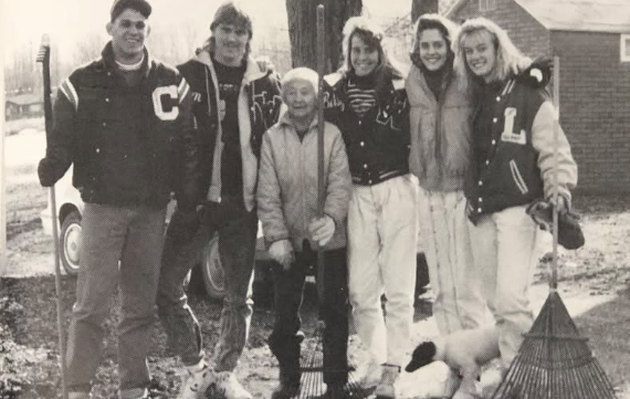 Black and white photo of five students standing with an older woman in a leaf-covered yard. The students are holding rakes and smiling at the camera.