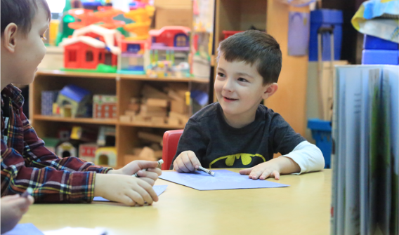 Two young boys sitting at a table coloring on paper and smiling at each other while in the classroom.