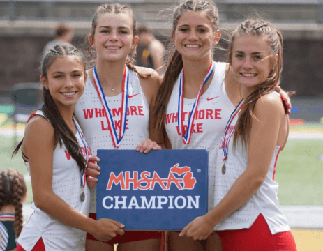 A group of girls in athletic outfits smiling at the camera while holding up a trophy they won for Girls Cross Country.