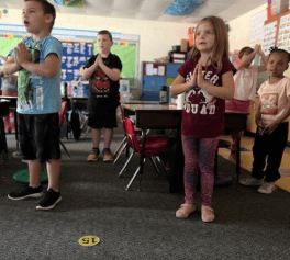 A teacher hugs a young female student in the classroom  while they smile at the camera.