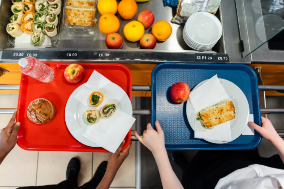 Two lunch trays being moved along the lunch line. The red tray has a water bottle, apple, muffin, and a plate with sandwich rolls. The blue tray has an apple and a plate with a sandwich.