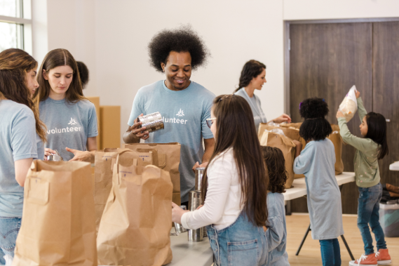 Employees and children gathered around tables with brown paper grocery bags filled with food.