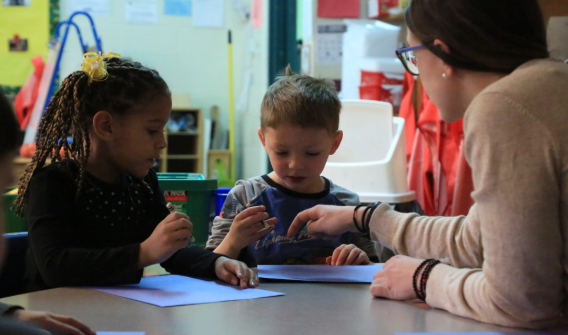 A teacher sitting at a table with a young boy and a young girl. The young children have a paper in front of them and the teacher is pointing at the paper while teaching the students.
